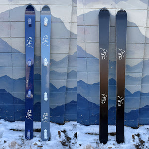 Split image of two hand painted shot skis, leaning against a wall with a mountain mural. Both have the Aria logo with the top of the skis painted blue and the bottom painted black.
