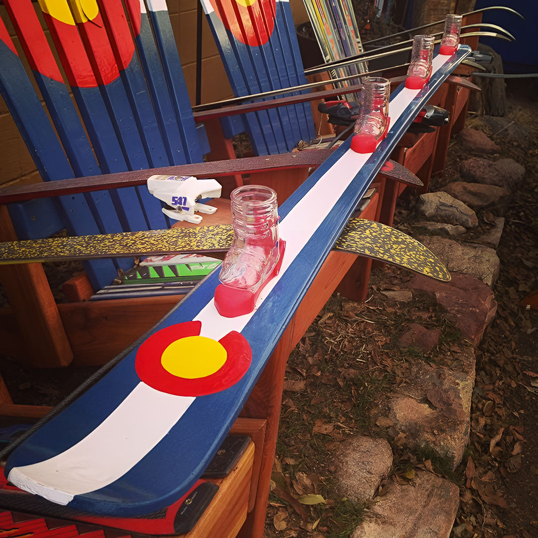 Hand painted 4 person shot ski with Colorado flag design on a wooden surface outdoors.
