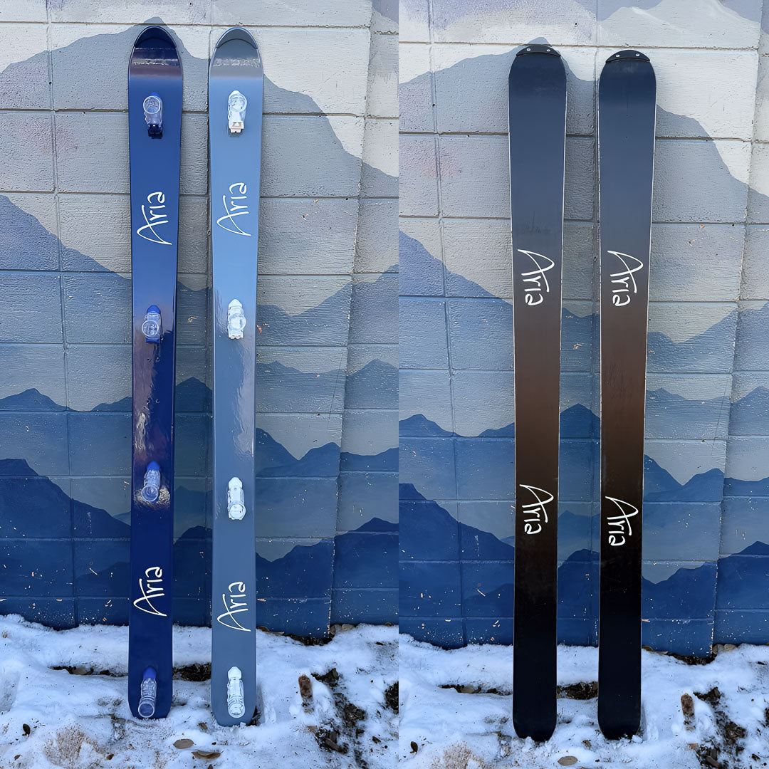 Split image of two hand painted shot skis, leaning against a wall with a mountain mural. Both have the Aria logo with the top of the skis painted blue and the bottom painted black.